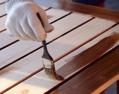 Hand with a brush paints a wooden surface with brown paint, close up